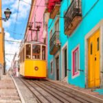 Yellow typical tram on a street with colorful houses and flowers on the balconies - Bica Elevator going down the hill of Chiado in Lisbon, Portugal.